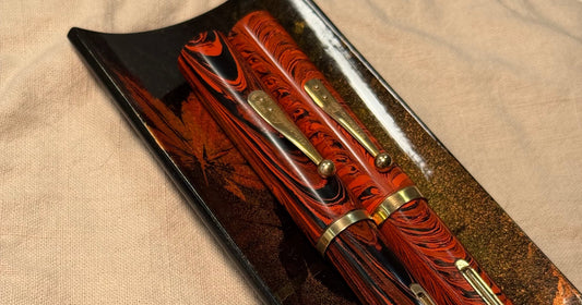 aesthetic photo of two waterman 58 ripple and woodgrain fountain pens resting on a maki-e urushi pen tray that is placed on top of a beige tea cloth with a drawing of blue flowers.